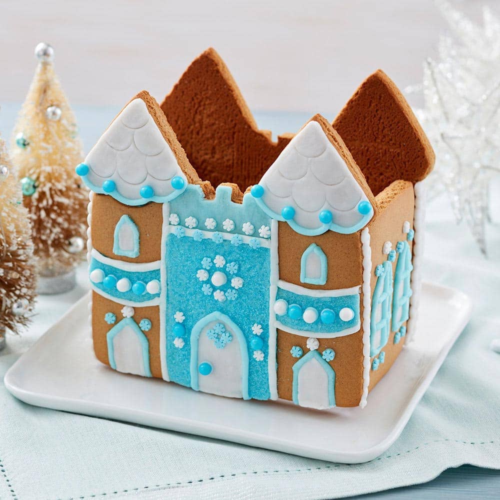Shimmer-Sparkle-Gingerbread-Castle Gingerbread castle with a blue front and white roof. White doors and windows, outlined with blue icing. White and blue round sprinkles decorate the front and sides of the castle.