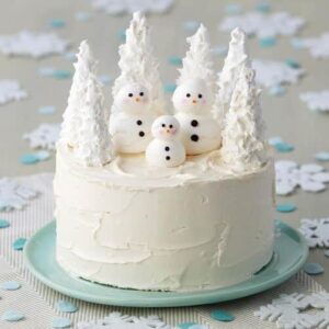 Round, white cake with three meringue snowmen with white trees made from ice cream cones behind them.