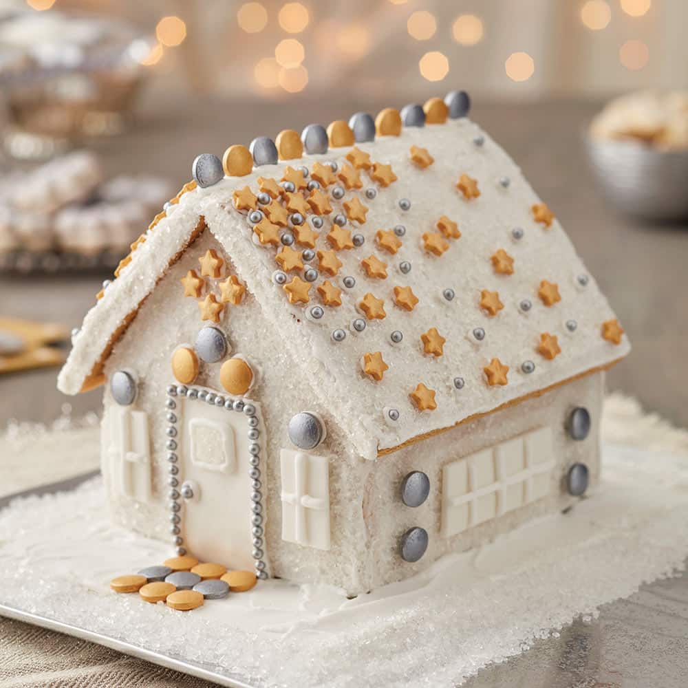 White, sparkly gingerbread house with gold star spinkle decorations and silver and gold dots lining the top of the roof and around the front door