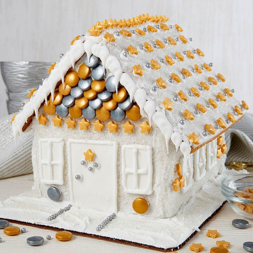 sparkle-gingerbread-house Sparkly white gingerbread house with gold star sprinkles lining the roof. Gold and silver circular sprinkles cover the front of the house above the door with gold stars in a row below.