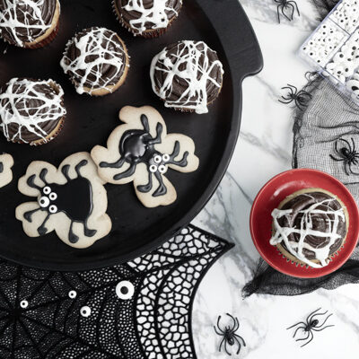 Vanilla cupcake with chocolate frosting with marshmallow spiderwebs surrounded by spider shaped sugar cookies